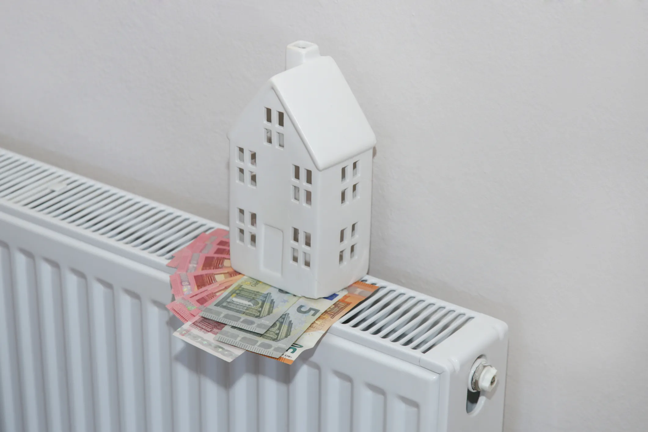 A small, white miniature house sits on a radiator with euro currency bills fanned out next to it, symbolizing heating costs.