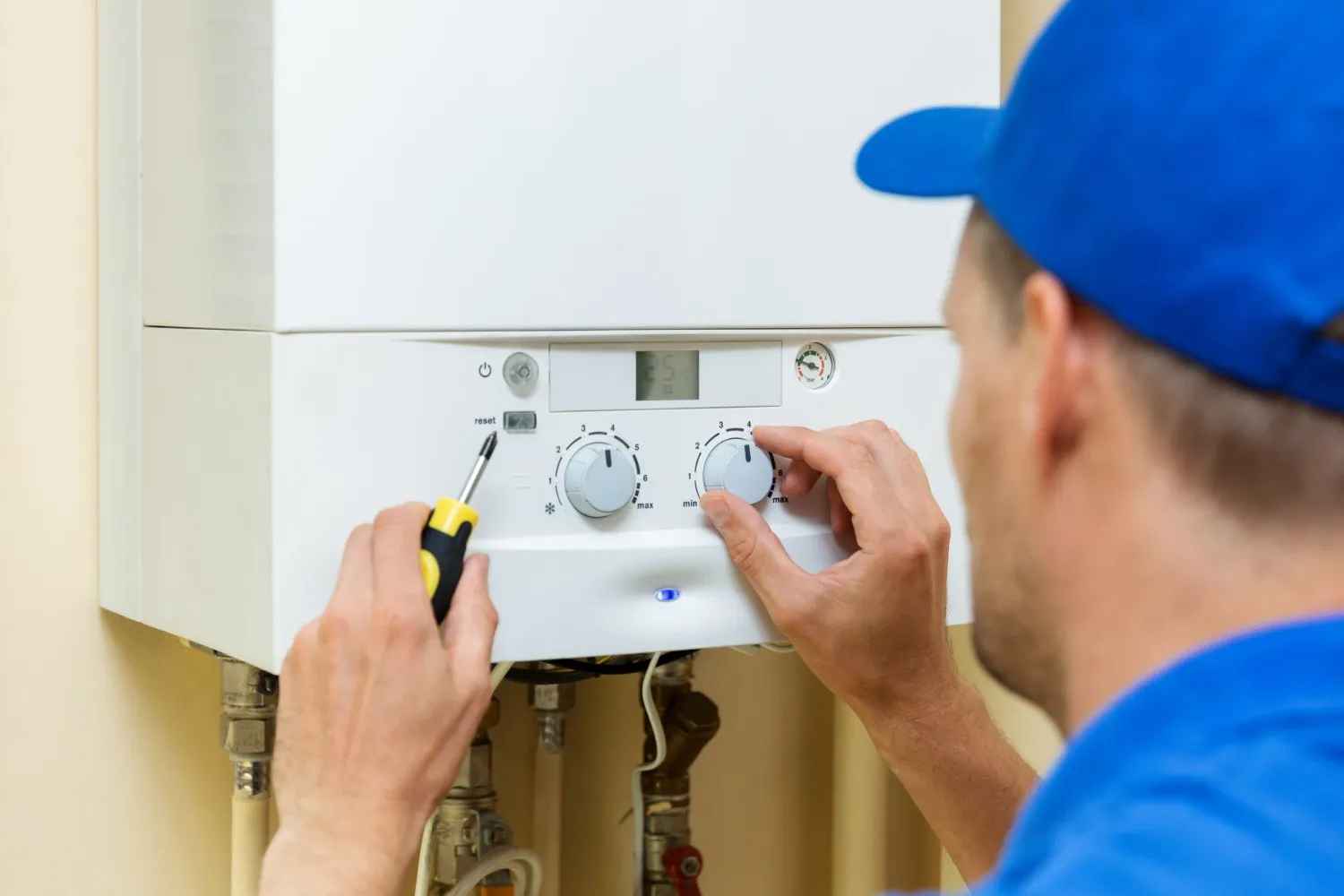  A service technician, wearing a blue shirt and baseball cap, is adjusting the controls on a white wall-mounted boiler or furnace. He is turning a temperature or pressure dial with his left hand, and holding a screwdriver near a 'reset' button with his right hand. The control panel has an LED screen and other dials for settings.