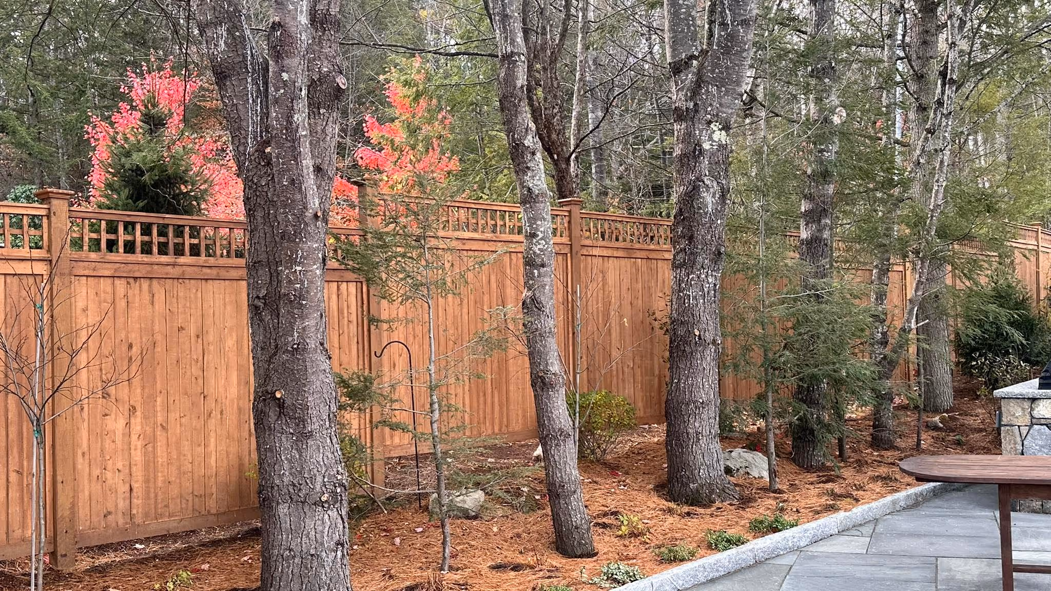 Tall wooden backyard fence with lattice top behind trees and a stone patio with a wooden table.