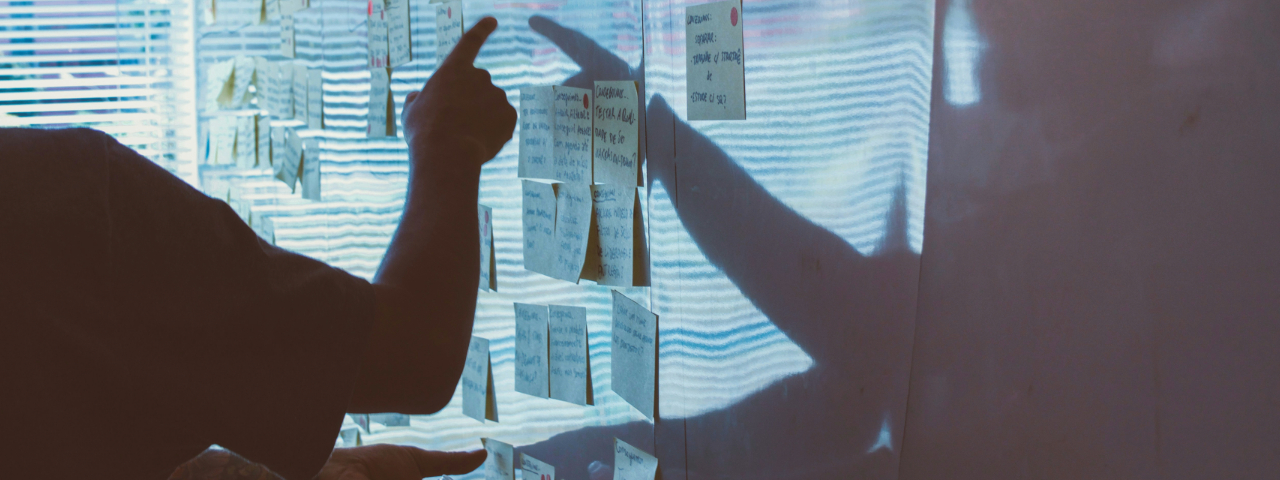 Person pointing at sticky notes on a whiteboard during a planning or brainstorming session, with natural light and shadows adding depth.