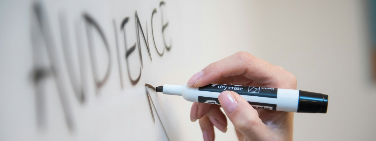 Hand writing the word 'AUDIENCE' on a whiteboard with a black dry erase marker.