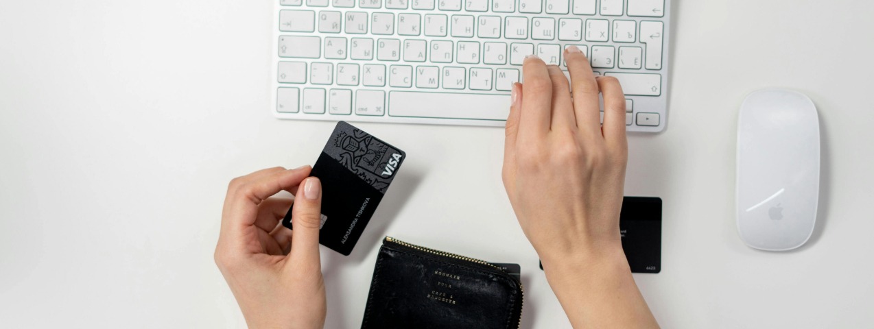 Person holding a credit card near a keyboard, with a black wallet and a white Apple mouse on a white desk.