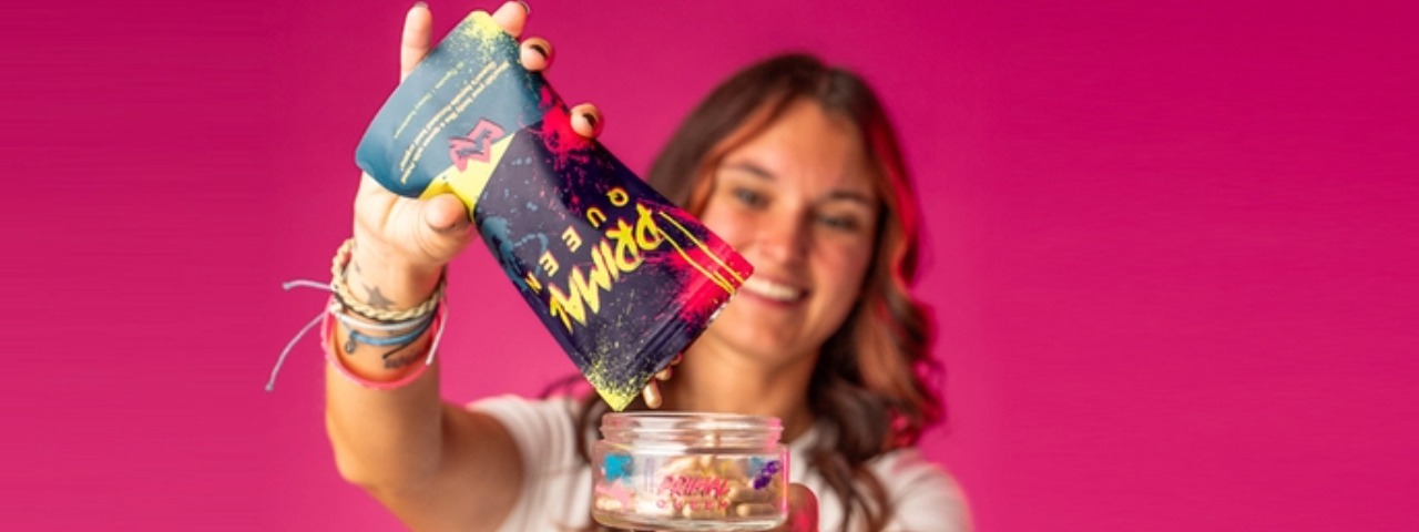 Woman pouring Primal Queen supplements into a jar against a pink background, showcasing product use and brand experience