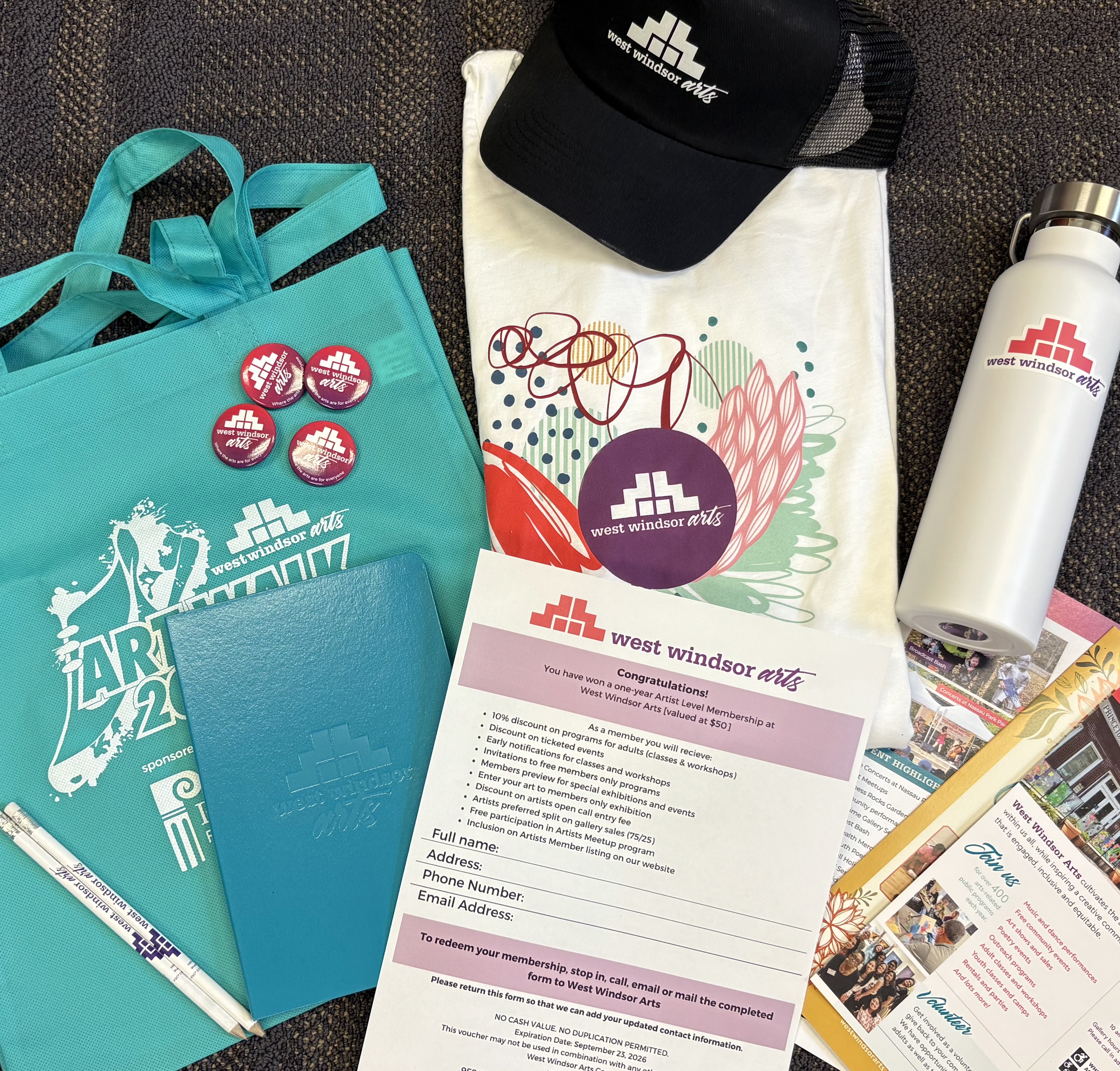 A collection of promotional items including a tote bag, t-shirt, water bottle, buttons, notebook, and membership form for West Windsor Arts.