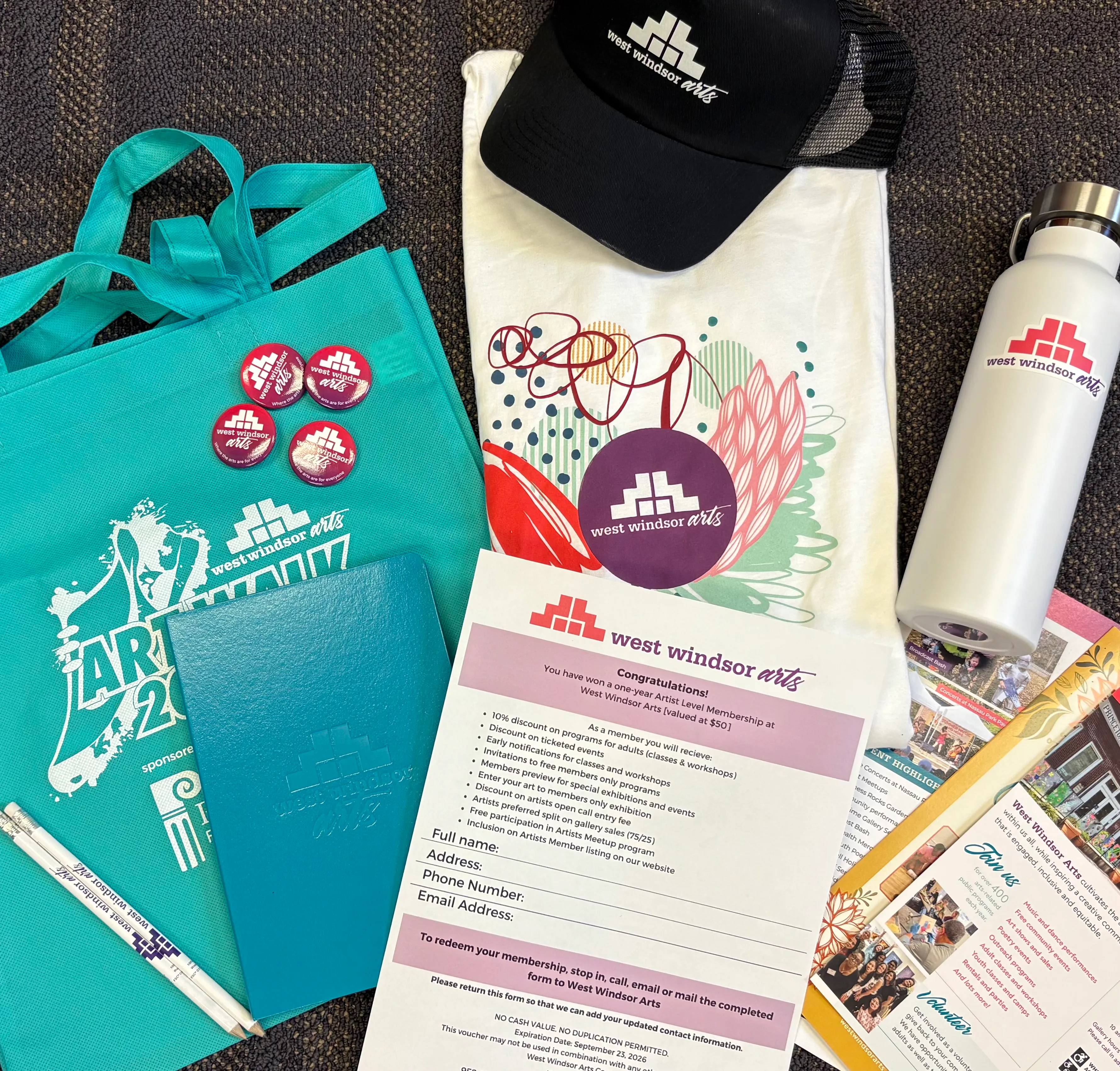 A collection of promotional items including a tote bag, t-shirt, water bottle, buttons, notebook, and membership form for West Windsor Arts.