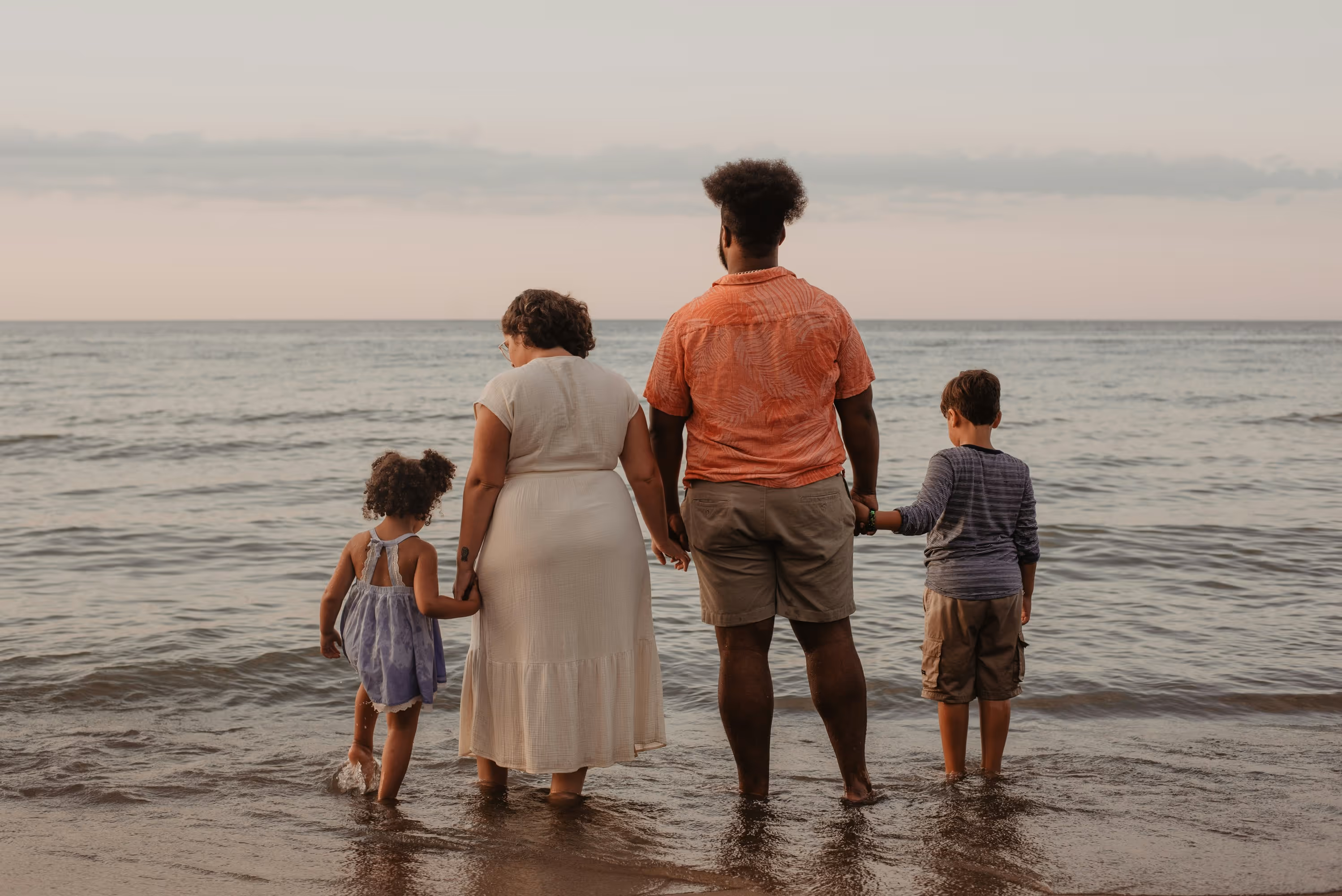 A family of four stands together in the shallow ocean waters at sunset, holding hands and gazing out at the horizon. The warm, golden hues of the setting sun reflect off the gentle waves, creating a serene and loving moment by the beach.
