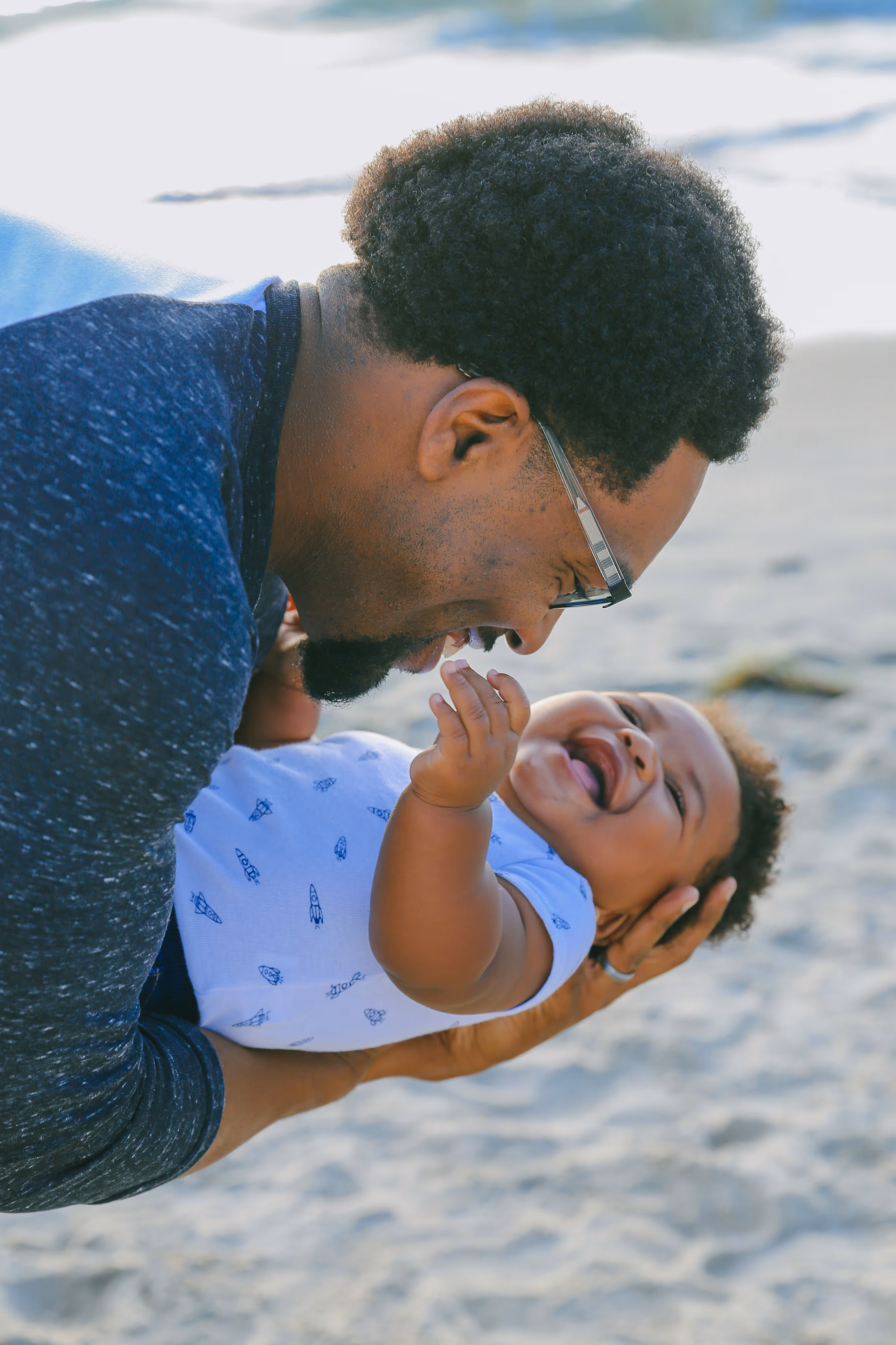 A father holds his child in his arms while playing on the beach. The child laughs with joy as the ocean waves roll in behind them, and the golden sunlight casts a warm glow over the sandy shore.