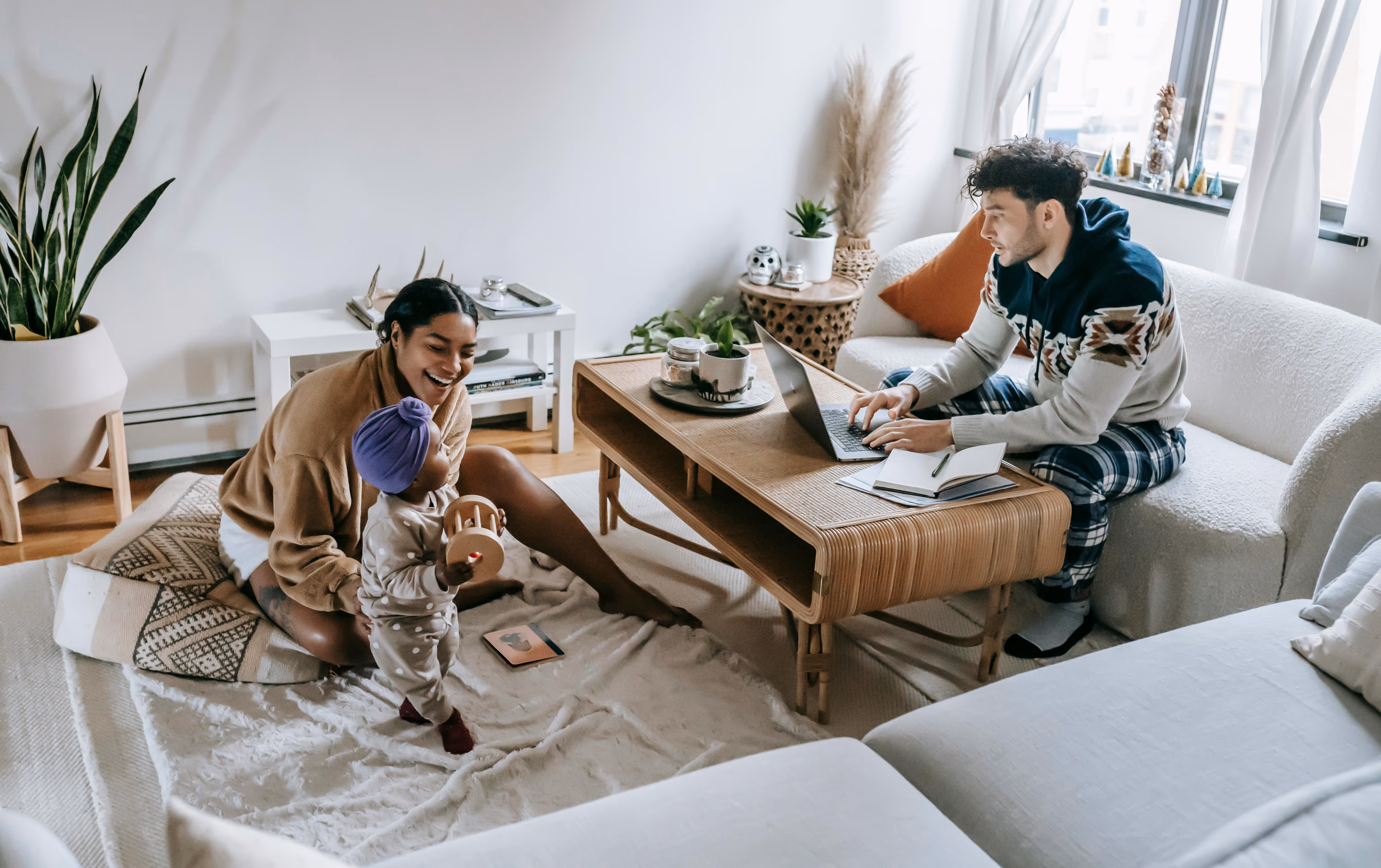 A young interracial family in a cozy living room; the father sits on the couch working on a laptop at the coffee table, while the mother sits on the floor playing with their toddler. Soft natural light fills the space, creating a warm and relaxed atmosphere.