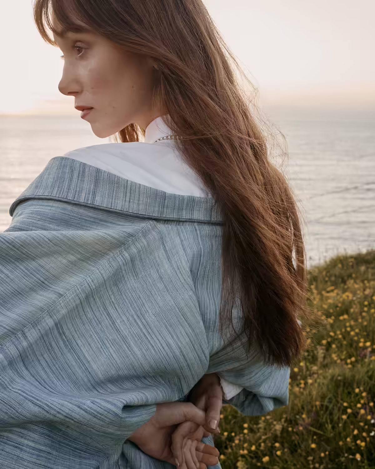 Woman with long brown hair wrapped in a blue shawl, standing near a grassy cliff overlooking the ocean at sunset.