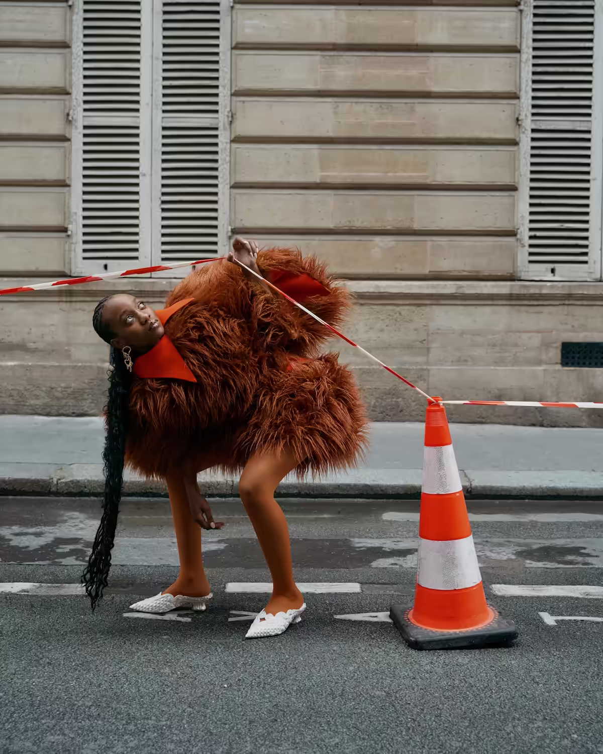 Woman in a fluffy brown coat and white woven shoes bending backward while holding red and white caution tape attached to a traffic cone on a city street.