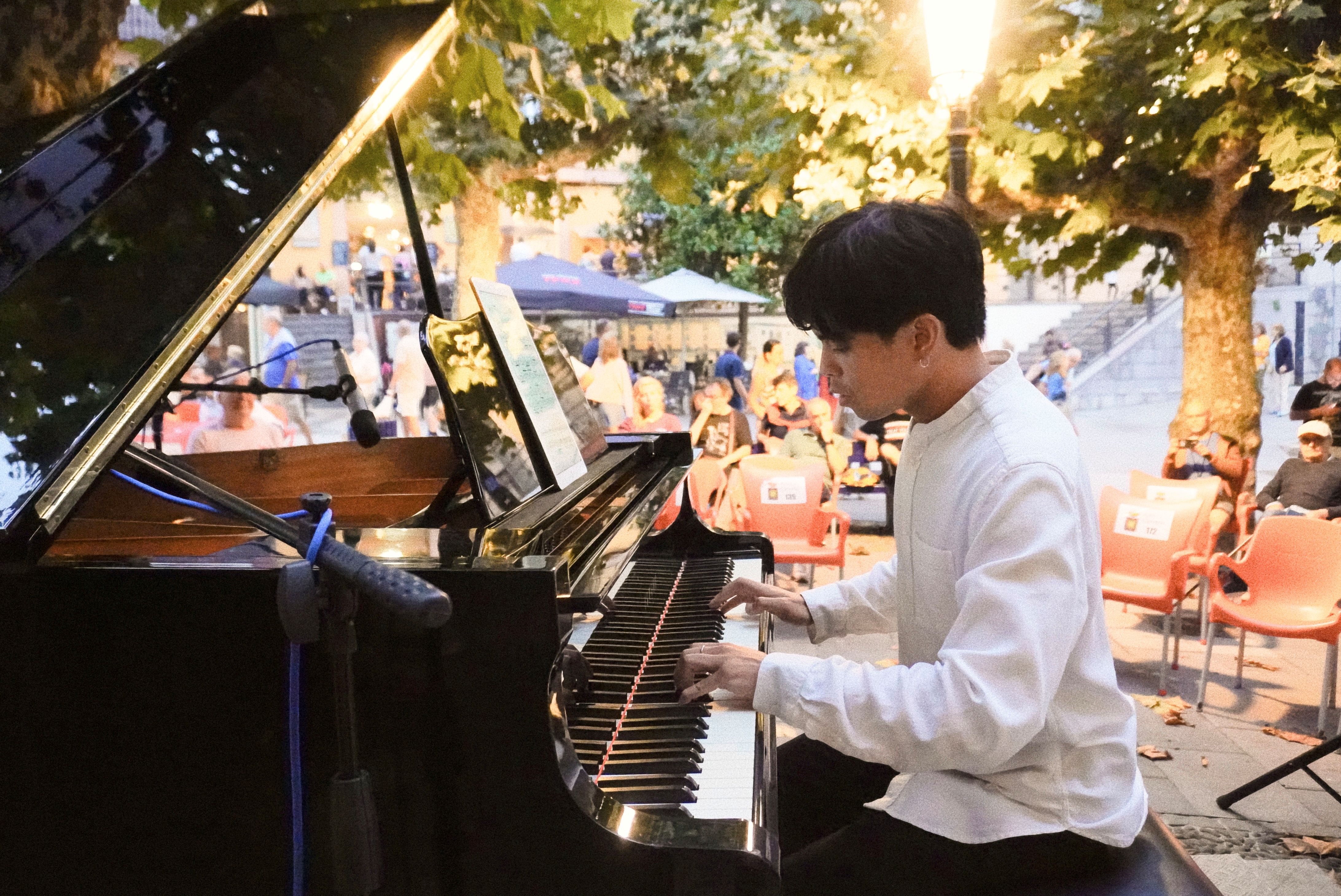 Young man in white shirt playing piano outdoors at an evening event with seated audience.