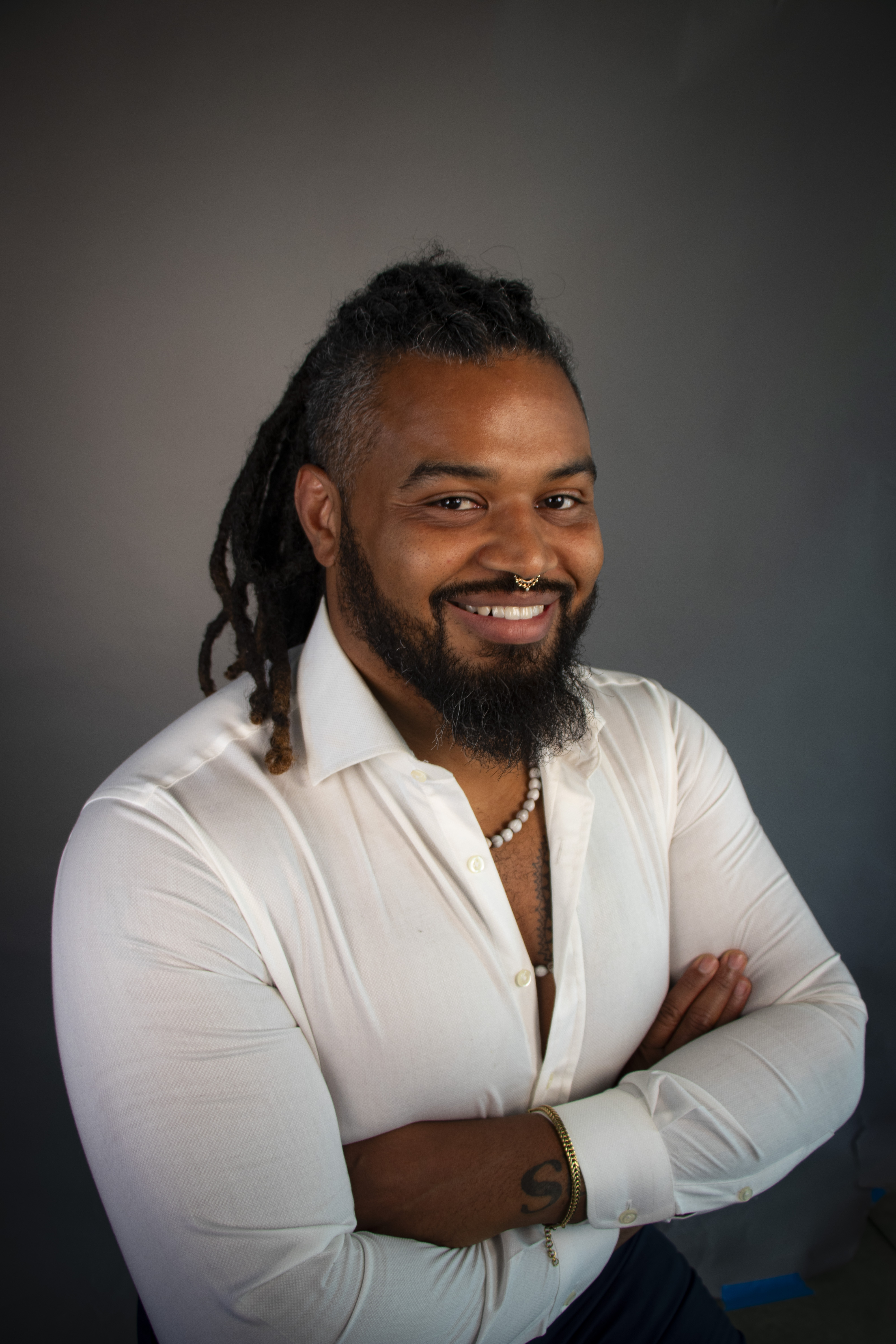 Smiling man with dreadlocks, beard, and septum piercing wearing a white shirt and pearl necklace, arms crossed.