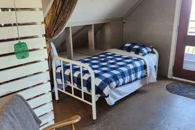 Small attic bedroom with a single bed covered in black and white checkered bedding, a wooden door leading outside, near a door with a window and a gray chair in the foreground.