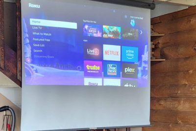 Roku streaming interface projected on a white screen in a wooden room, showing options like Home, Live TV, Netflix, Prime Video, and Plex.