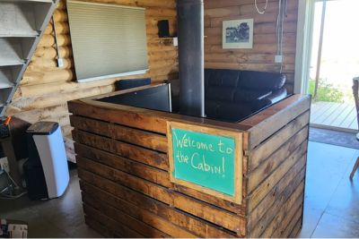 Cozy cabin interior with a wood-burning stove enclosed by a wooden fire wall with a green sign saying 'Welcome to the Cabin!' and a couch in the background.