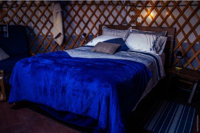 Cozy bed with blue blanket and pillows inside a yurt featuring lattice wooden walls and soft lighting.