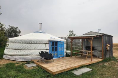 White yurt with blue door next to a wooden deck and a small shed with picnic table under an overcast sky.