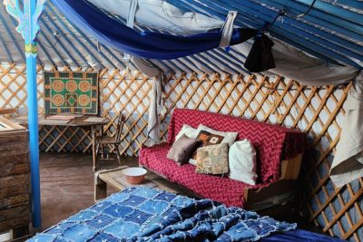 Interior of a yurt featuring a blue bedspread, a red couch with multiple decorative pillows, and a small table with a colorful quilt hanging behind it.