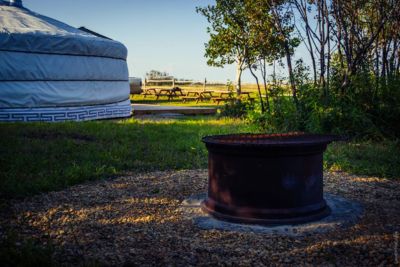 Metal fire pit on a gravel surface with a yurt and picnic tables in the background surrounded by grass and trees.