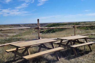 Two wooden picnic tables on grass overlooking a scenic landscape with a wooden fence and a partly cloudy blue sky.