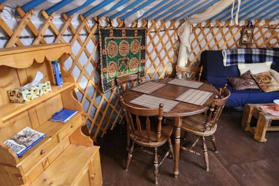 Cozy interior of a yurt with wooden furniture including a round dining table with four chairs, a blue couch with pillows, and a wooden shelf.