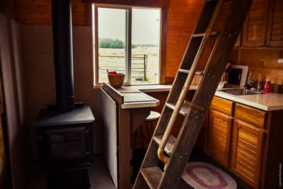 Cozy wooden cabin kitchen with a wood-burning stove, ladder, stool, and window showing an outdoor view.