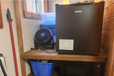 Black mini fridge on a wooden shelf with a microwave next to a small black fan and a blue storage bin underneath.