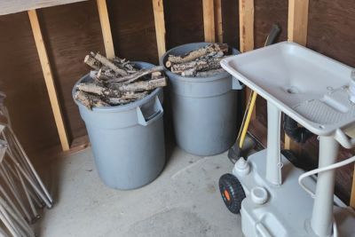 Two gray bins filled with cut firewood inside a wooden outhouse next to a portable sink.