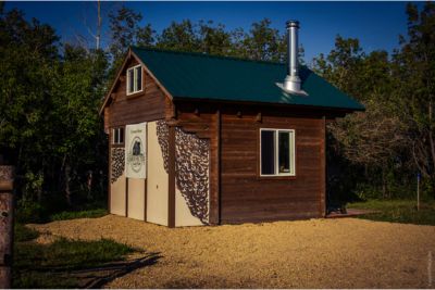 Small wooden cabin with green metal roof and chimney surrounded by trees and gravel path.