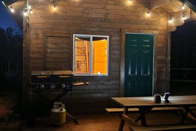 Wooden cabin exterior at night with lights, a lit window, green door, picnic table, and a grill.