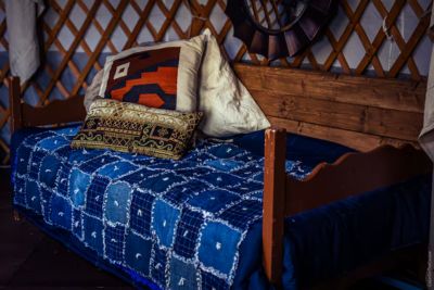 Wooden bed with blue patchwork quilt and patterned throw pillows inside a yurt.