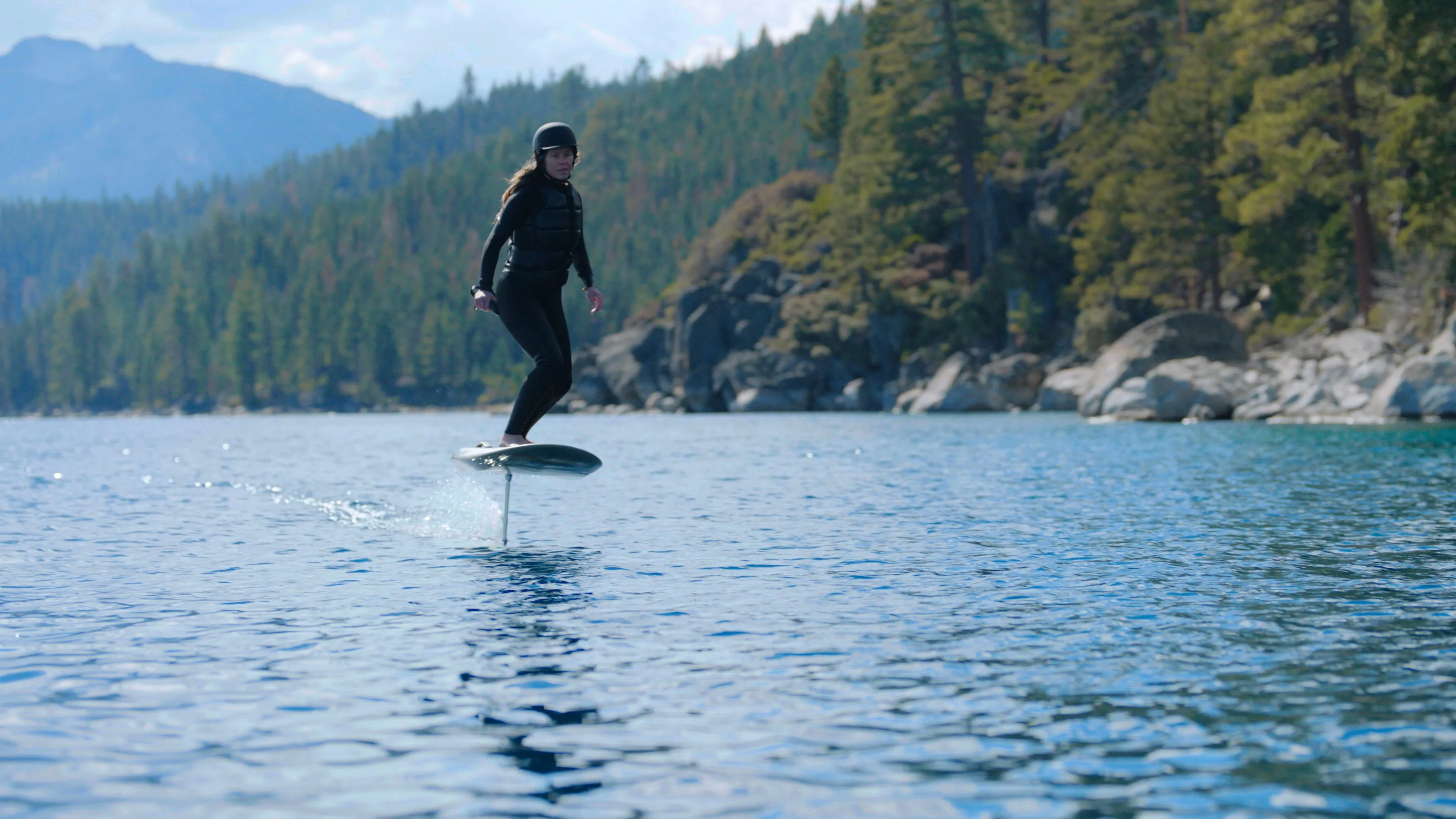 Rider in a wetsuit and helmet flying an eFoil above Lake Tahoe, with pine-covered shoreline behind.