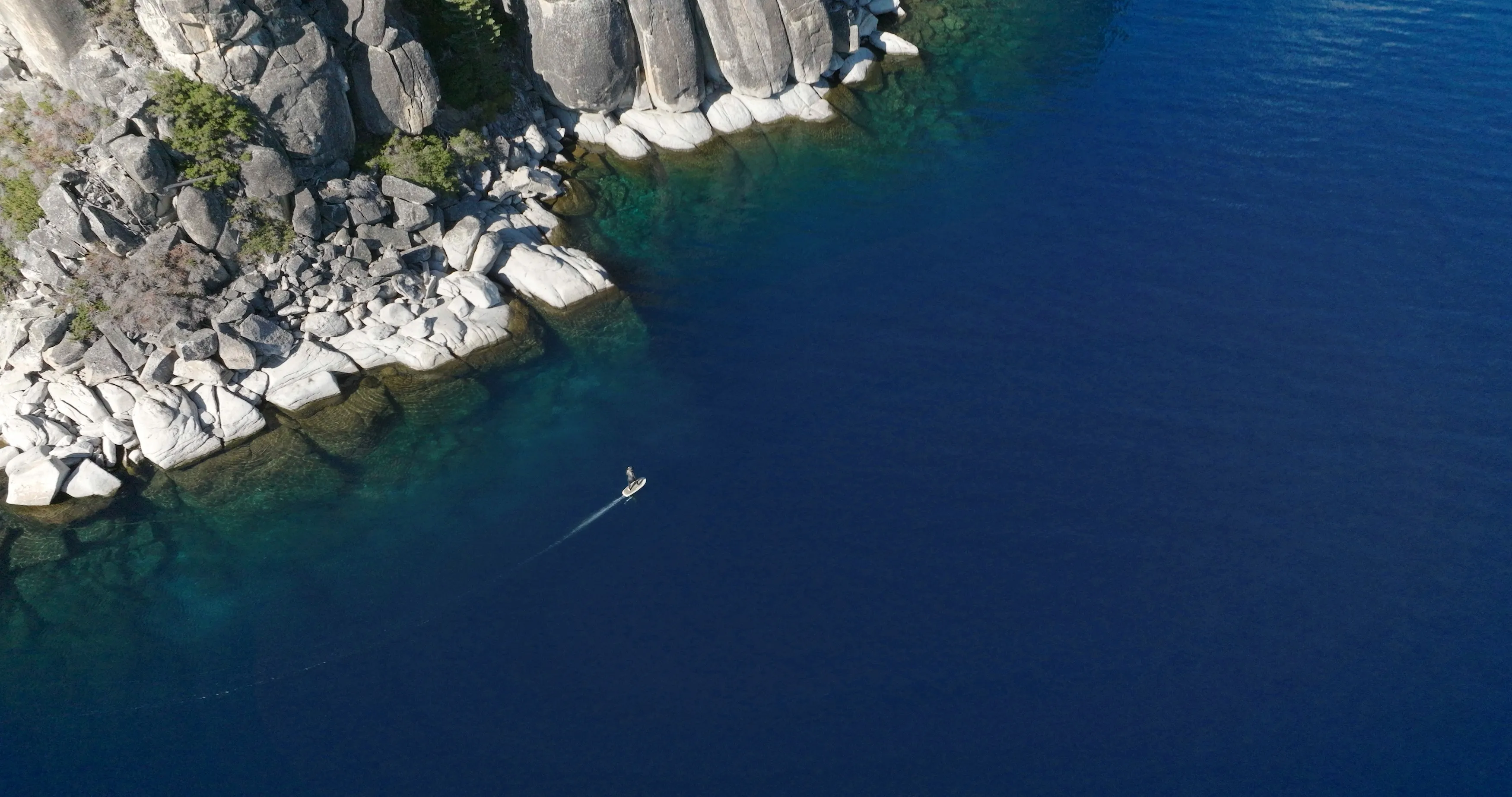 Aerial view of an eFoil rider gliding past granite boulders along Lake Tahoe's clear blue shoreline.
