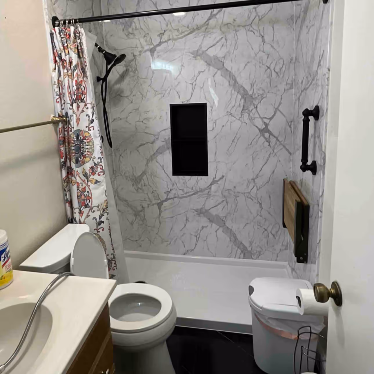 Bathroom with marble-patterned shower walls, a white toilet, a floral shower curtain, wooden cabinet sink, and trash can with toilet paper holder on the door.