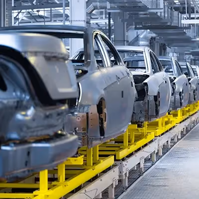 Unfinished car bodies move along a yellow conveyor on an automotive assembly line inside a modern factory, showing multiple vehicle frames in sequence during manufacturing.