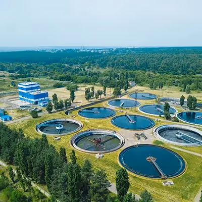 Aerial view of a wastewater treatment facility with multiple circular treatment tanks surrounded by greenery, trees, and open landscape, showing an organized layout of water processing infrastructure.