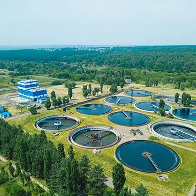 Aerial view of a wastewater treatment facility with multiple circular treatment tanks surrounded by greenery, trees, and open landscape, showing an organized layout of water processing infrastructure.