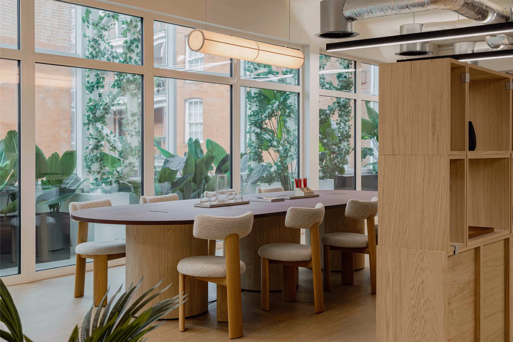 Modern office with six swivel chairs around a long table, desk lamps, plants, and large windows on the left.