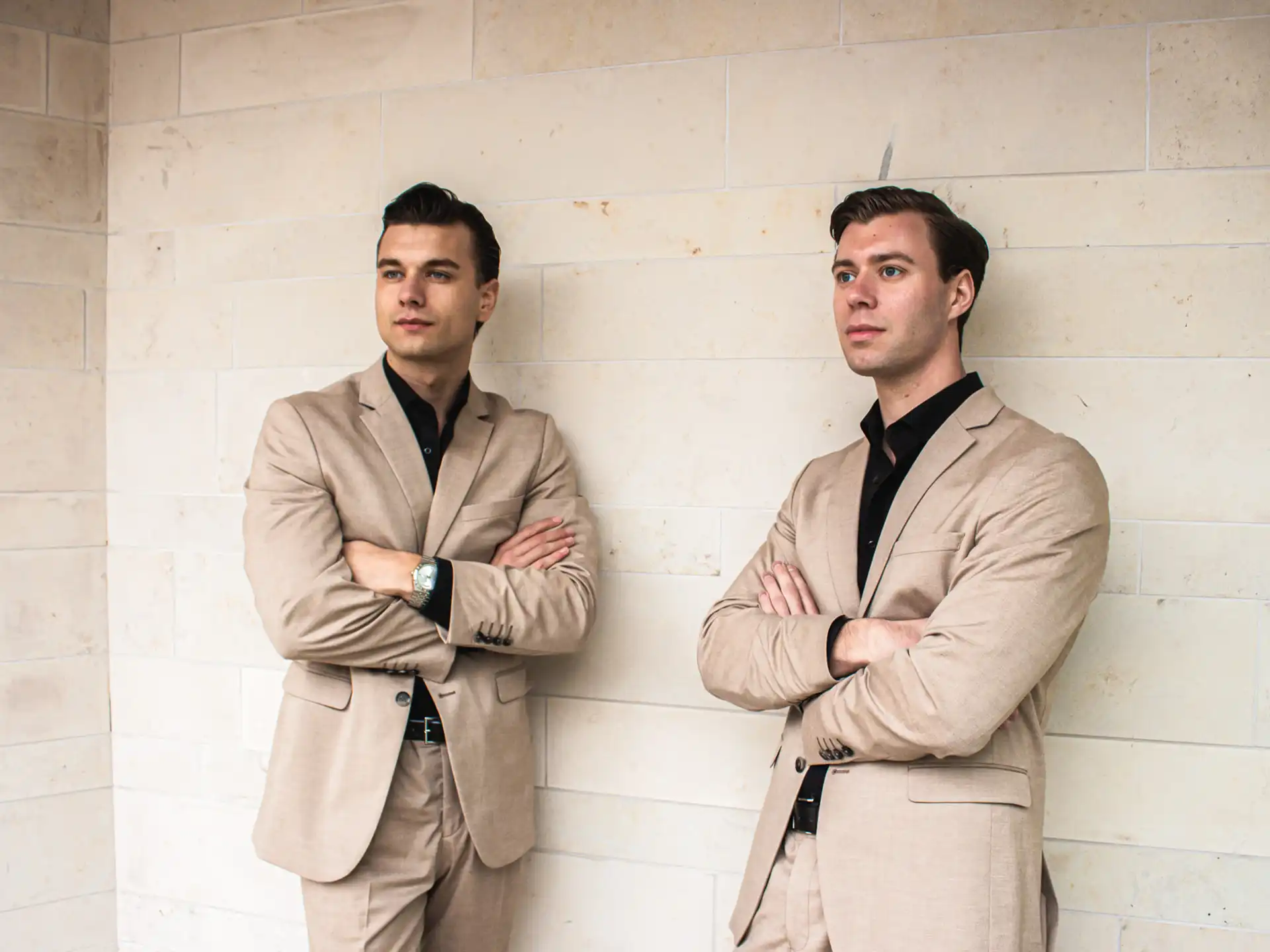 Two men wearing beige suits and black shirts standing with arms crossed against a light stone wall.