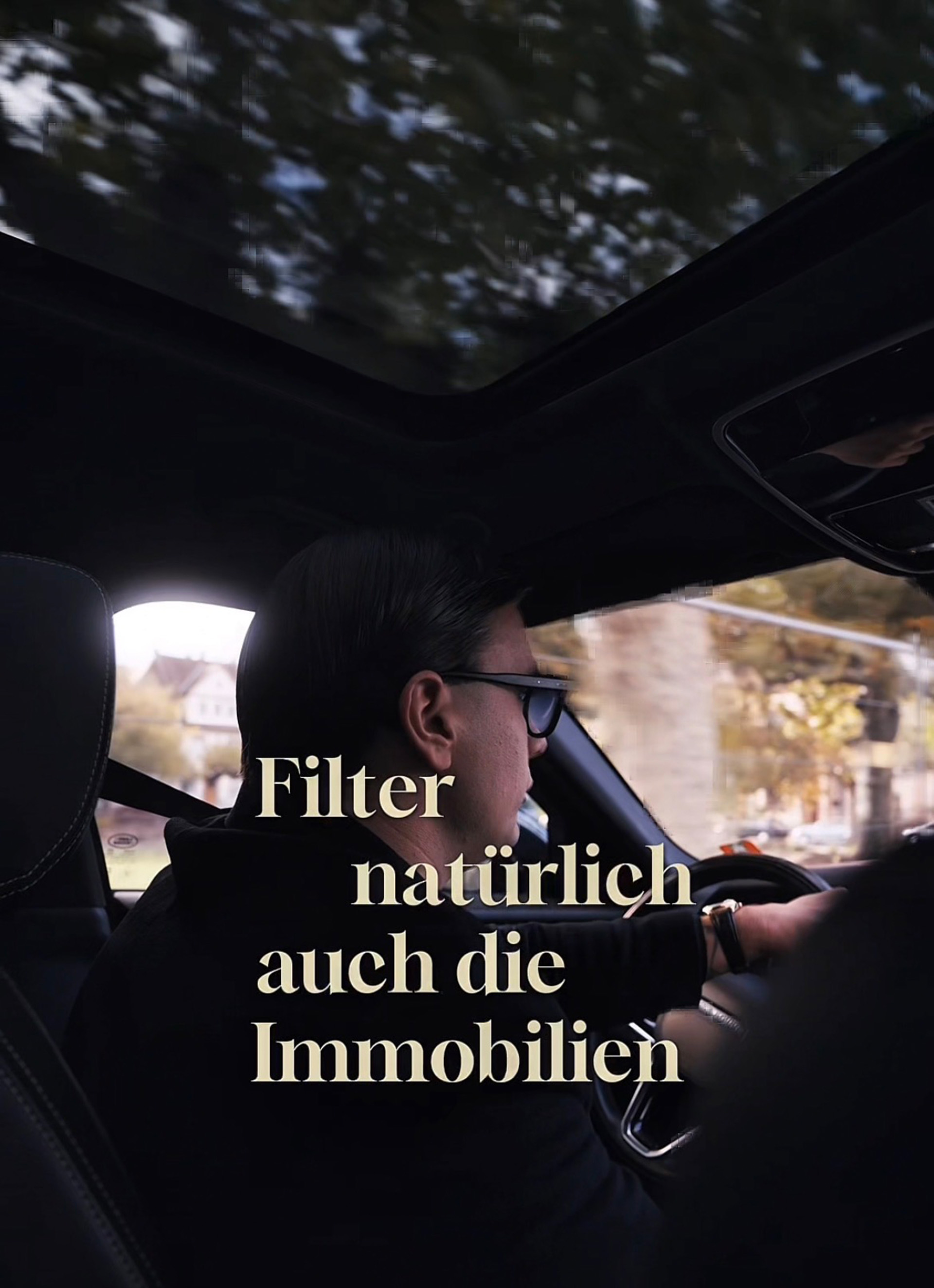 Man wearing glasses driving a car with trees visible through the windows and a sunroof above.