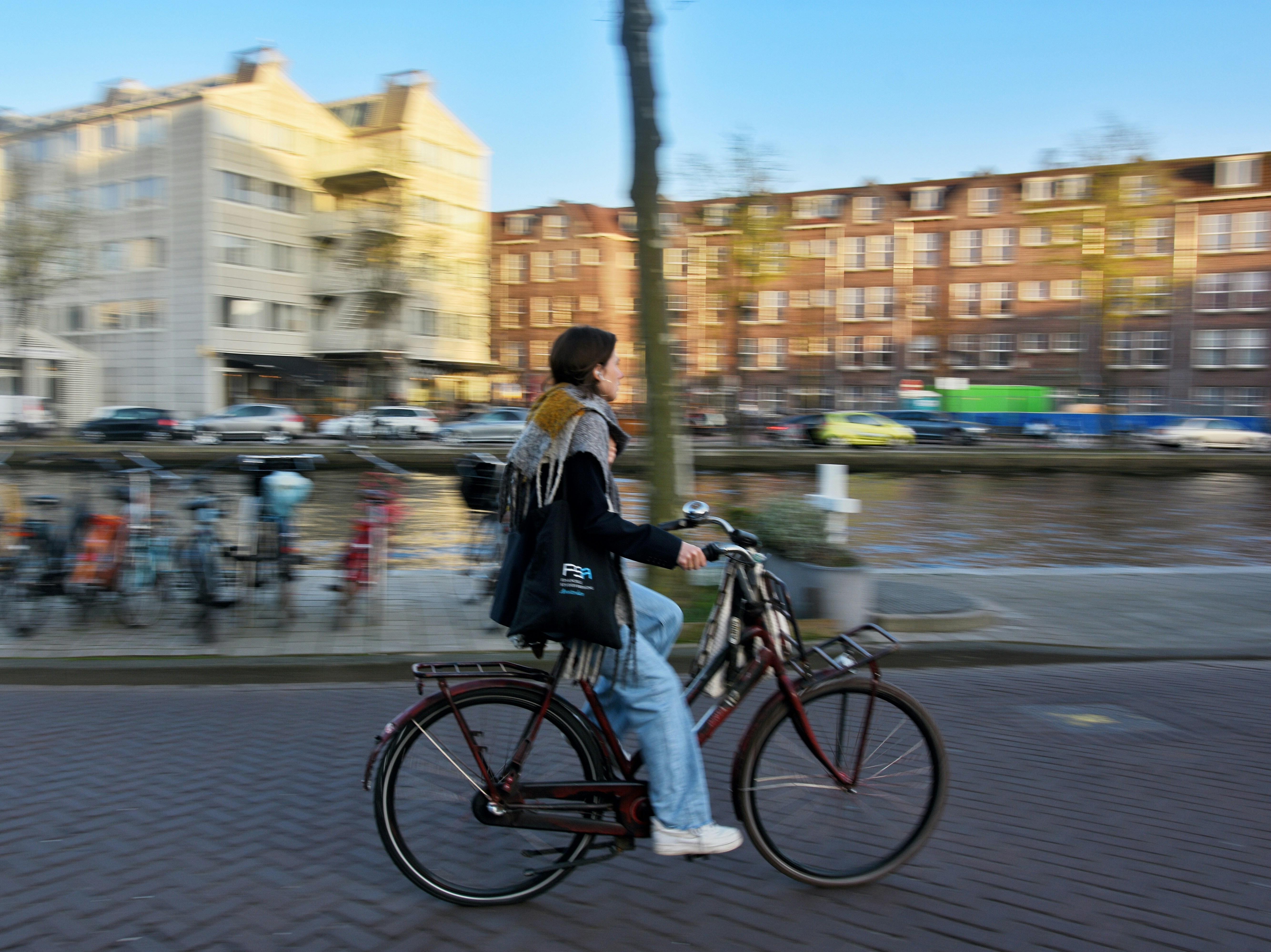 En kvinde cykler langs en kanal i byen, omgivet af bygninger og parkerede cykler - relevant i diskussionen om regler for, om man må køre fuld på cykel i trafikken.