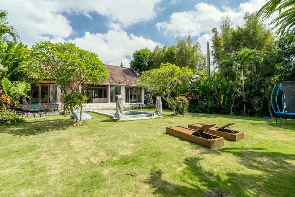 Balinese tiled-roof villa viewed across a landscaped lawn with a rectangular pool, carved stone statues and a trampoline – Villa Paminna, a 4-bedroom villa in Tandeg, Berawa, Bali