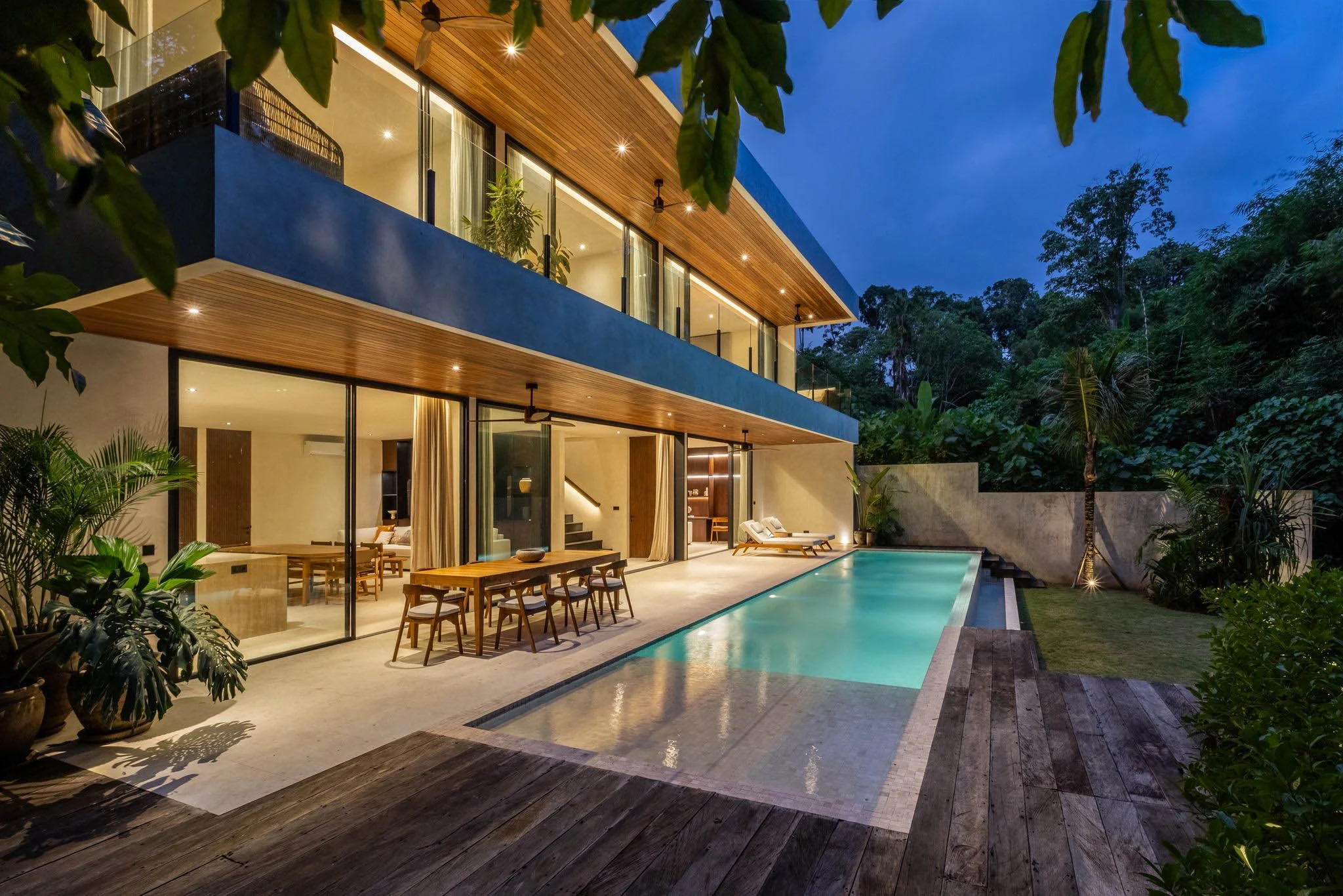 Two-story modern villa lit at dusk with a wood-clad upper balcony, glass-walled dining area, and a long pool running alongside the terrace – Villa Belanza, a 4-bedroom villa in Tumbak Bayu, Pererenan, Bali