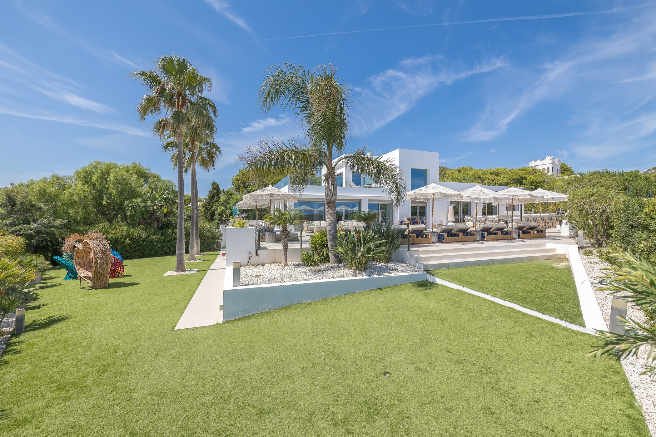 Modern white villa seen across a manicured lawn with tall palm trees and a shaded terrace lined with white-fringed umbrellas and daybeds – Villa Azura, a 4-bedroom villa