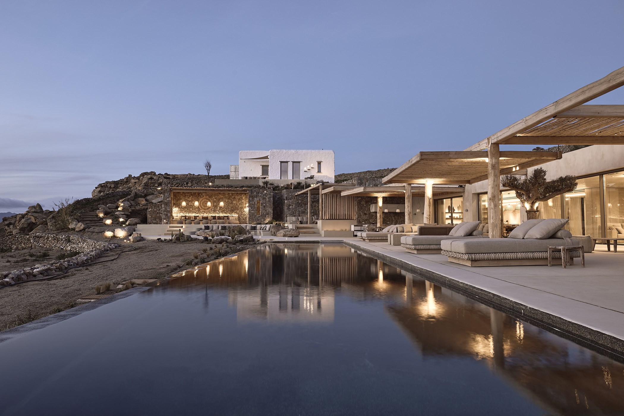 Stone-and-timber villa at dusk with a dark reflective infinity pool along a covered lounge terrace with pergola and daybeds – Prestige, a 7-bedroom villa