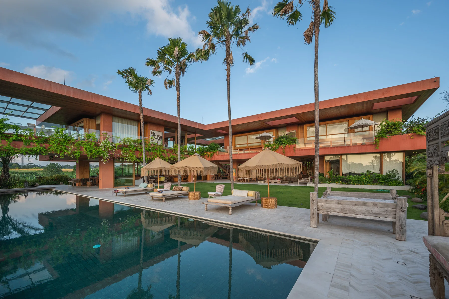 L-shaped two-story villa with rust-toned roofline and timber accents around a long reflecting pool lined with sun loungers, fringed umbrellas and palm trees – Villa Merida, a 6-bedroom villa in Tumbak Bayu, Pererenan, Bali
