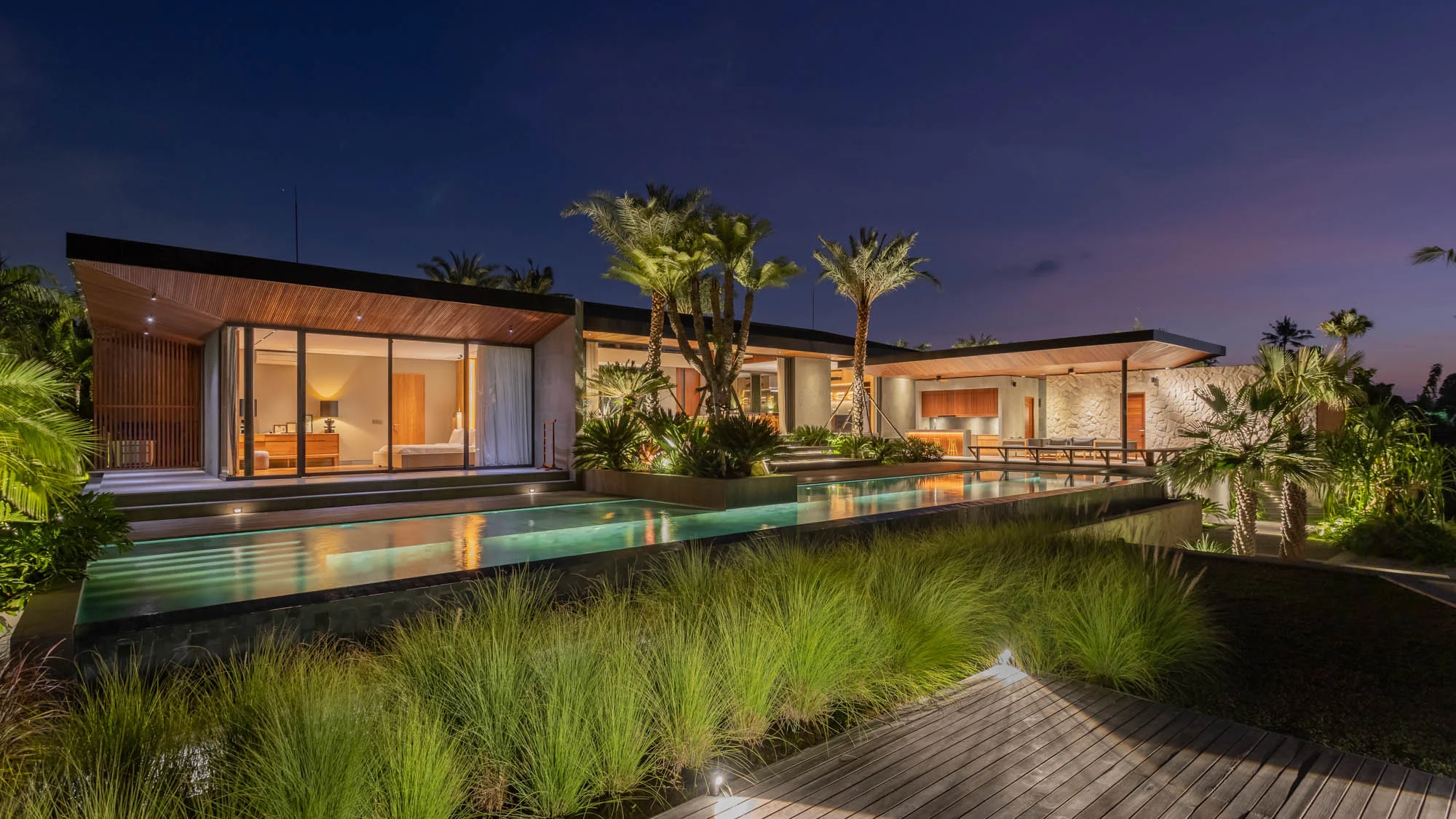 Single-story villa at night with a wood-lined overhanging roof, glass-walled lit bedroom and a long narrow pool running past ornamental grasses and palms – Villa Korya, a 4-bedroom villa in Nyambu, Kaba Kaba, Bali
