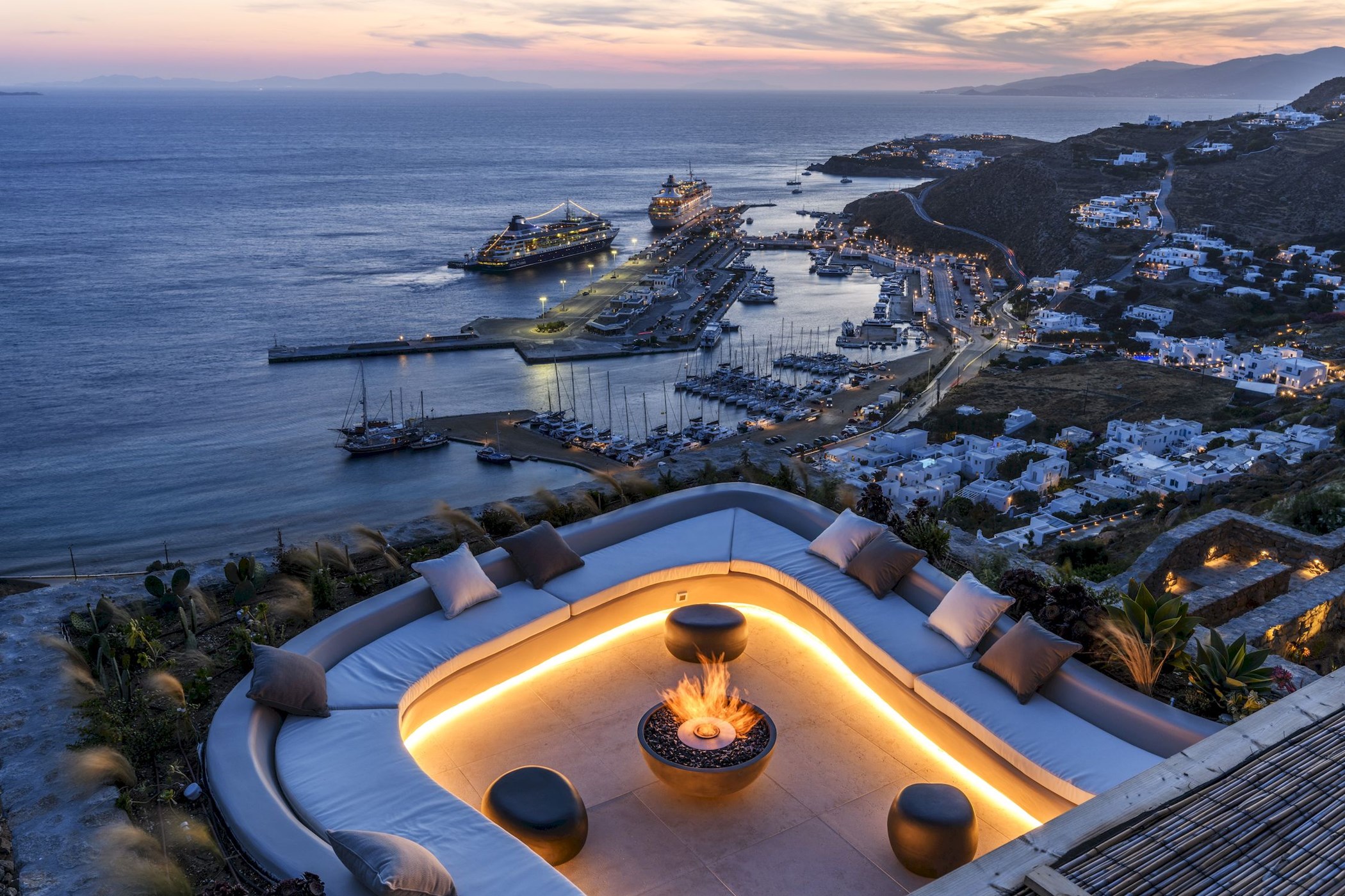 Rooftop lounge with curved sofa and fire pit overlooking a harbor town at dusk with cruise ships and moored yachts below – Paradise Escape, an 8-bedroom villa