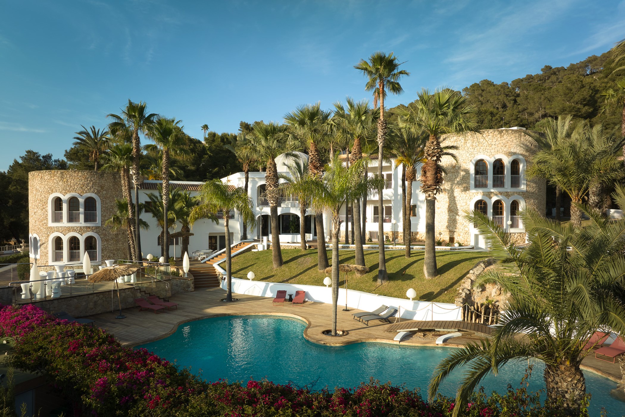 Ibiza-style stone-and-white villa with round turret towers among tall palm trees beside a free-form pool with wooden deck and pink bougainvillea in the foreground – Villa Amado, a 6-bedroom villa