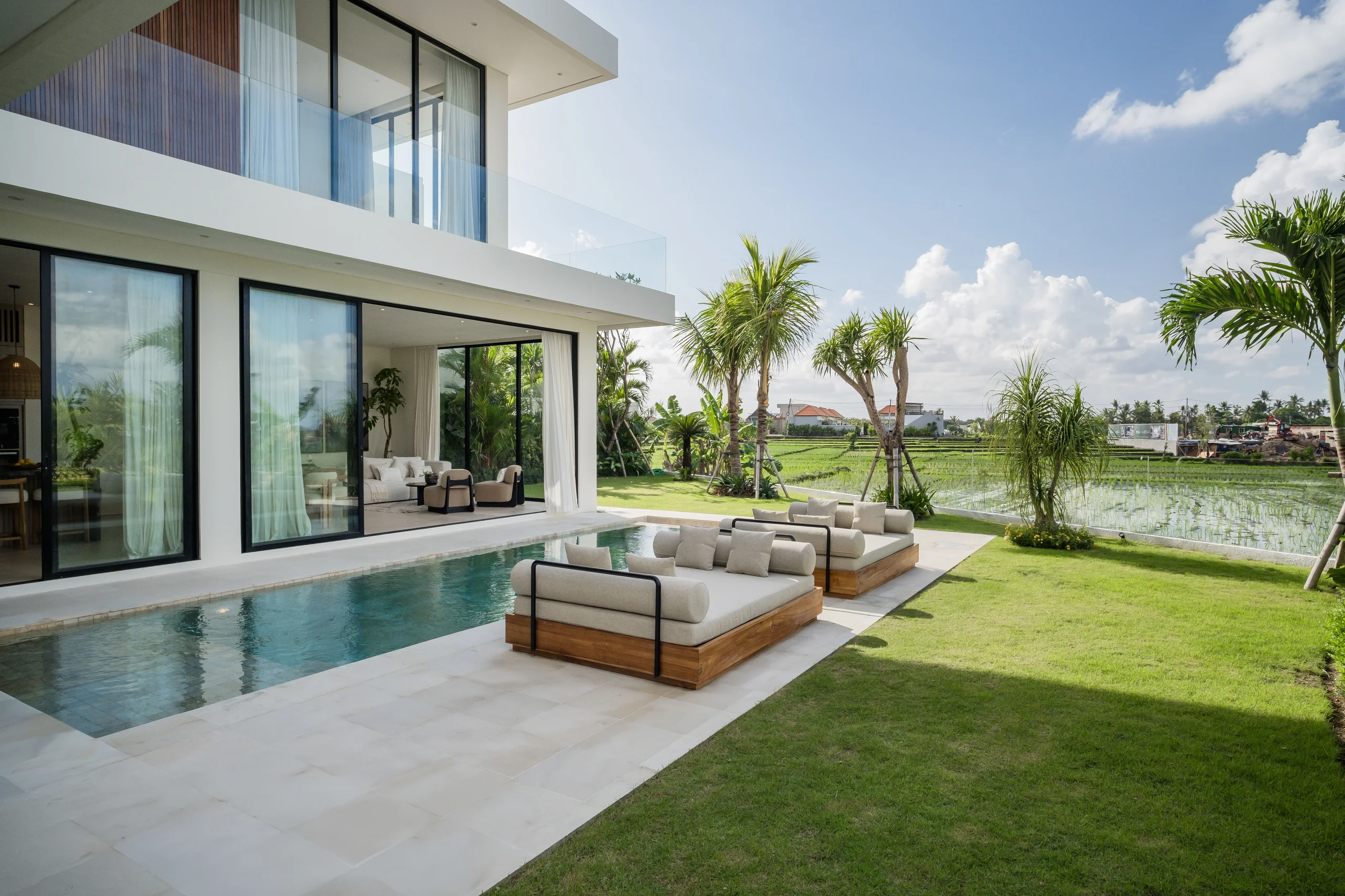 Modern white villa with a pool along a lawn edged by two large wood-framed daybeds, overlooking open rice paddy fields – Villa Mavala, a 4-bedroom villa in Seseh, Bali